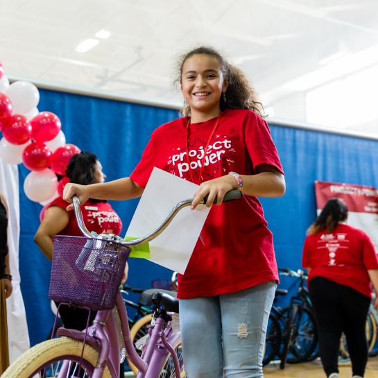 A young girl holds up a bike while wearing a project power shirt
