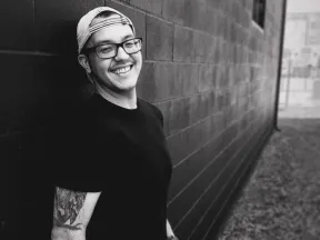 man with glasses and hat smiling in grayscale leaning against brick wall