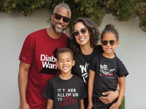 A family of four stands in front of a leafy green wall. The father wears a dark red t-shirt that says "Diabetes Warrior" in white letters. The mother wears a black t-shirt that says "We Fight" in white letters. The young son wears a black t-shirt with the words "It Takes All of Us to Stop Diabetes" written on it. The young daughter wears a black t-shirt with the words "We Fight" in white letters. All are smiling and wearing sunglasses.