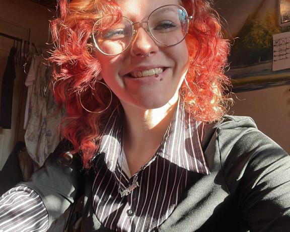 A selfie of a person with curly red hair and glasses smiling into the camera.