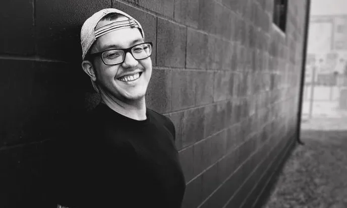 man with glasses and hat smiling in grayscale leaning against brick wall
