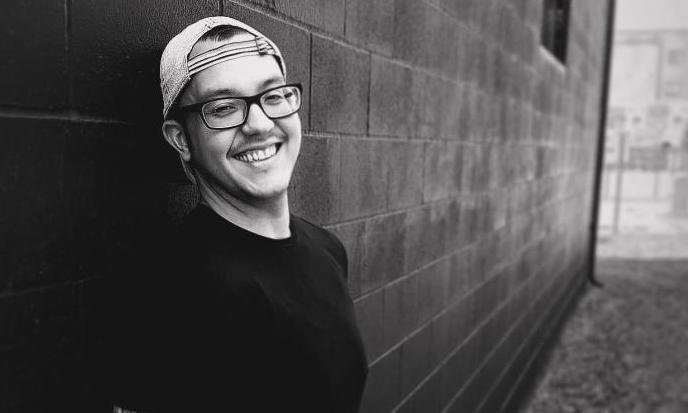 man with glasses and hat smiling in grayscale leaning against brick wall