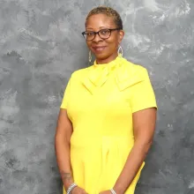 Photo of woman in yellow standing in front of gray background.