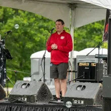Man in red sweatshirt and shorts speaks into a microphone on an outdoor stage.