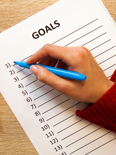 A hand holding a blue pen prepares to fill out a list of goals. 