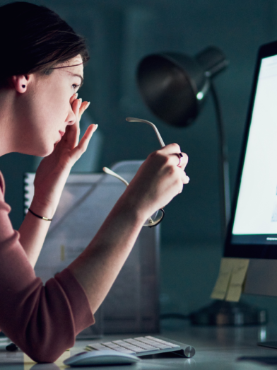 A woman sits in front of a large computer screen and rubs at her eyes